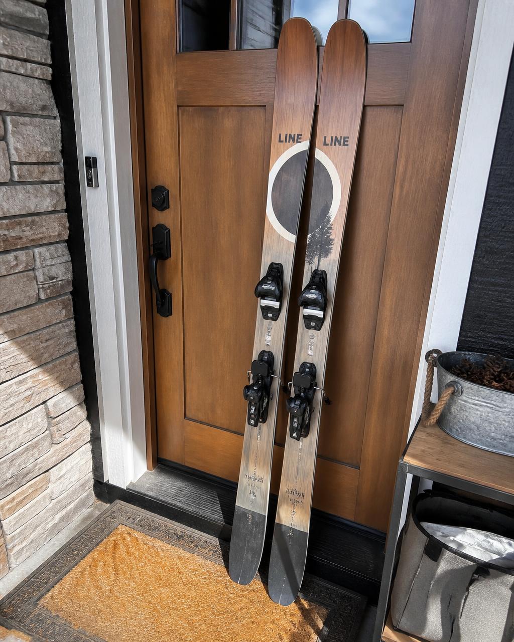 A pair of used all-mountain skis with bindings leaning against a wooden front door